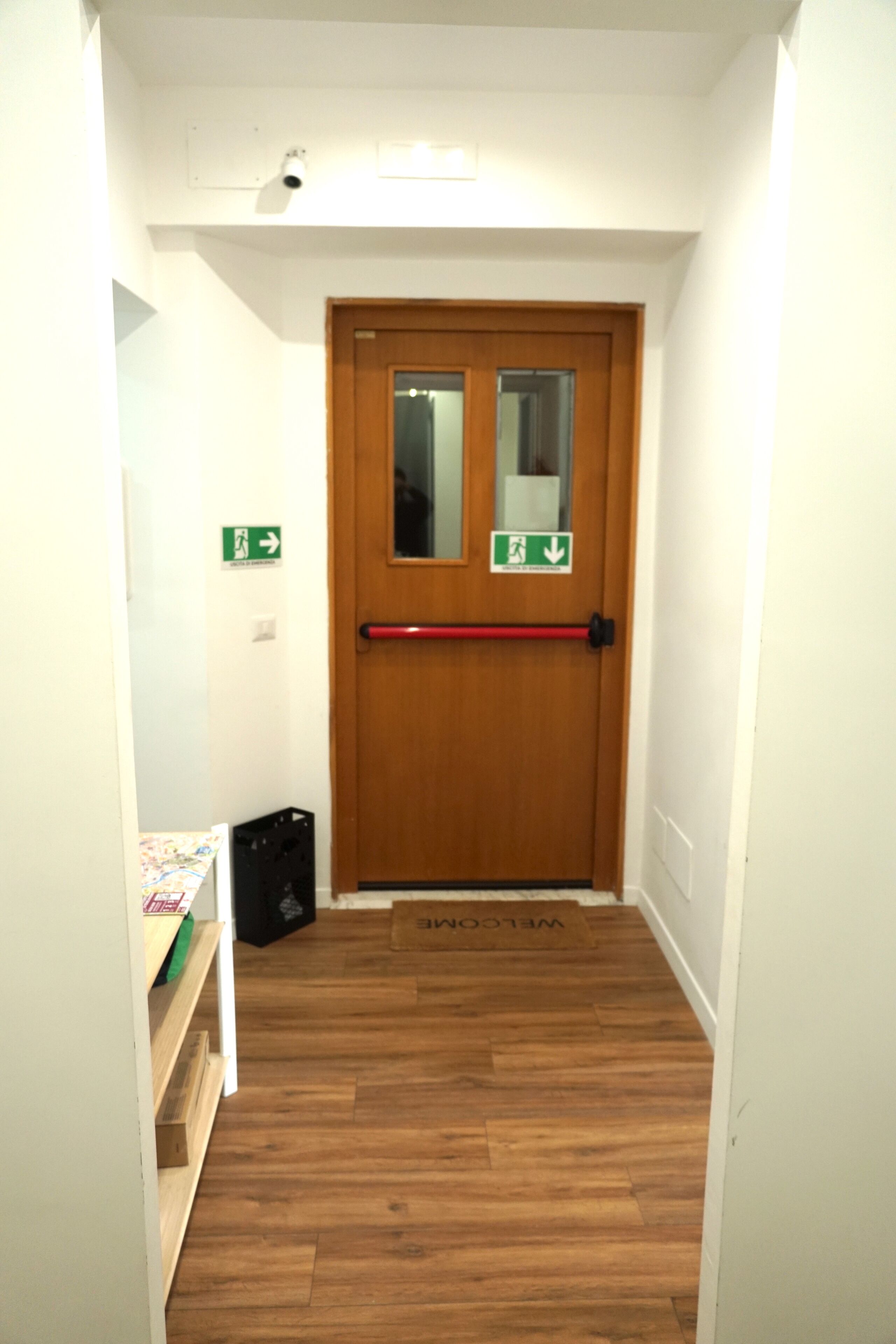 Een hal met een houten deur, een rode deurklink en een welkomstmat. Er hangen groene borden met richtingaanwijzingen aan de muur en een zwarte tas ligt op de vloer.