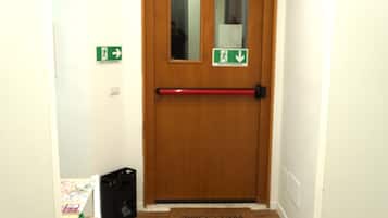 Een hal met een houten deur, een rode deurklink en een welkomstmat. Er hangen groene borden met richtingaanwijzingen aan de muur en een zwarte tas ligt op de vloer.