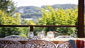Una mesa de madera con sillas blancas en un balcón, con vistas a un paisaje verde con árboles y colinas.