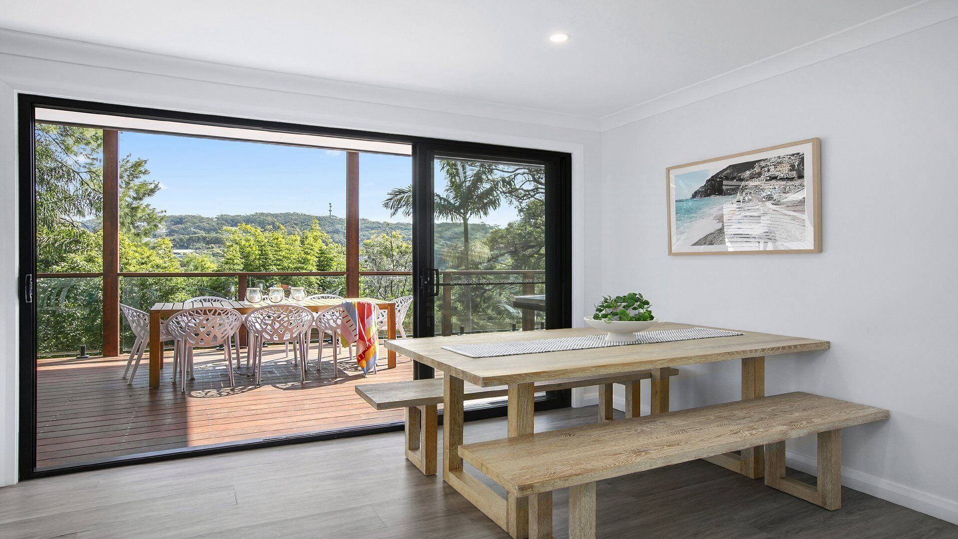 A room with a wooden table and bench, a balcony with chairs, and a framed picture on the wall.