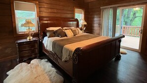 A wooden bedroom with a large bed, two bedside tables with lamps, a chandelier, and a view of the outdoors through a sliding glass door. - Charming 3,800 sq feet log house in Waupaca County.  (Ogdensburg)