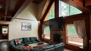 A cozy living room with a curved green leather sofa, a stone fireplace, and large windows offering a view of the outdoors. - Charming 3,800 sq feet log house in Waupaca County.  (Ogdensburg)