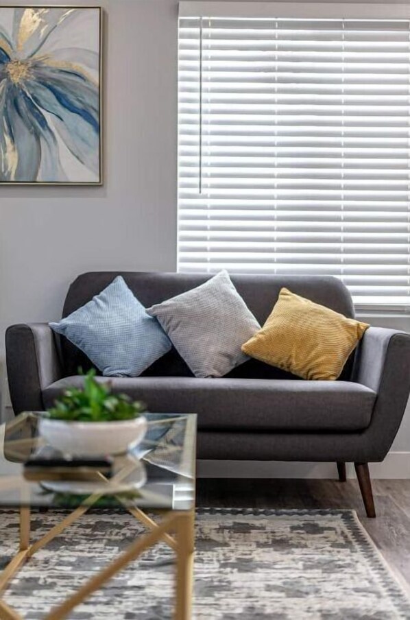 A modern living room with a grey sofa, three cushions, a glass coffee table with a plant, and a patterned rug. - Ocean Breeze, Newly Remodeled, Playroom (Chula Vista)