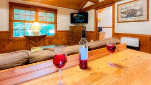Interior de madera con una mesa de madera, dos copas de vino, una botella de vino, un televisor y un cuadro en la pared.