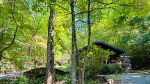 Una cabaña rústica con base de piedra y techo verde, rodeada de un bosque denso y un puente de piedra.