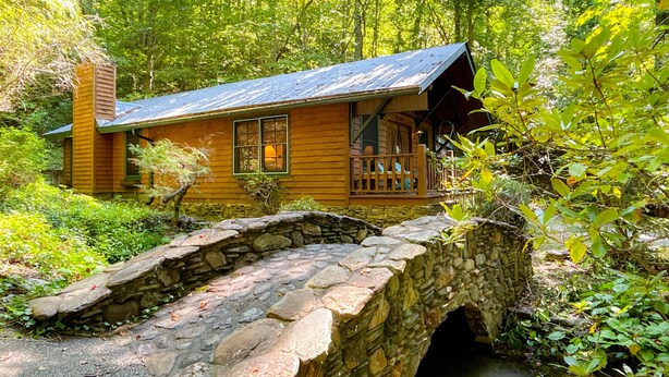 Una cabaña de madera rústica con un puente de piedra que lleva a un porche, rodeada de abundante vegetación.