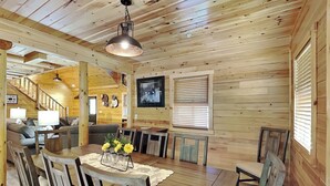 Ein Holzblockhaus-Interieur mit einem Esstisch, Stühlen, einer Kristallleuchtre, einem gerahmten Bild und einer Treppe, die in einen anderen Raum führt.