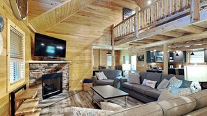 Ein gemütliches Blockhaus-Interieur mit Kamin aus Stein, Holzböden, einem Wohnzimmer mit Sofa und Couchtisch, einem an der Wand befestigten Fernseher und einem Balkon mit Holzbalkonen.