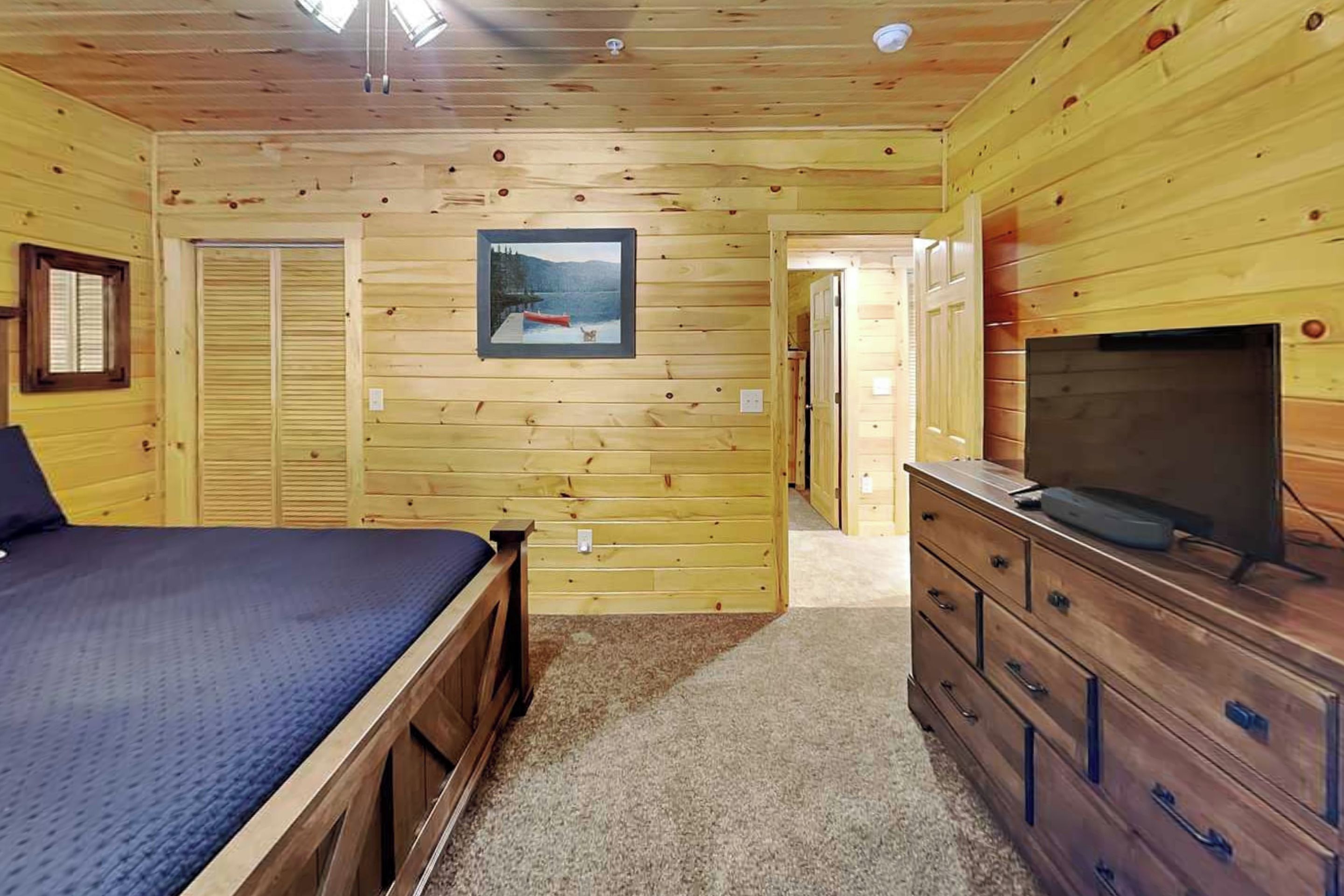 Une chambre rustique en bois, avec un lit, une commode, un téléviseur et un tableau encadré au mur.