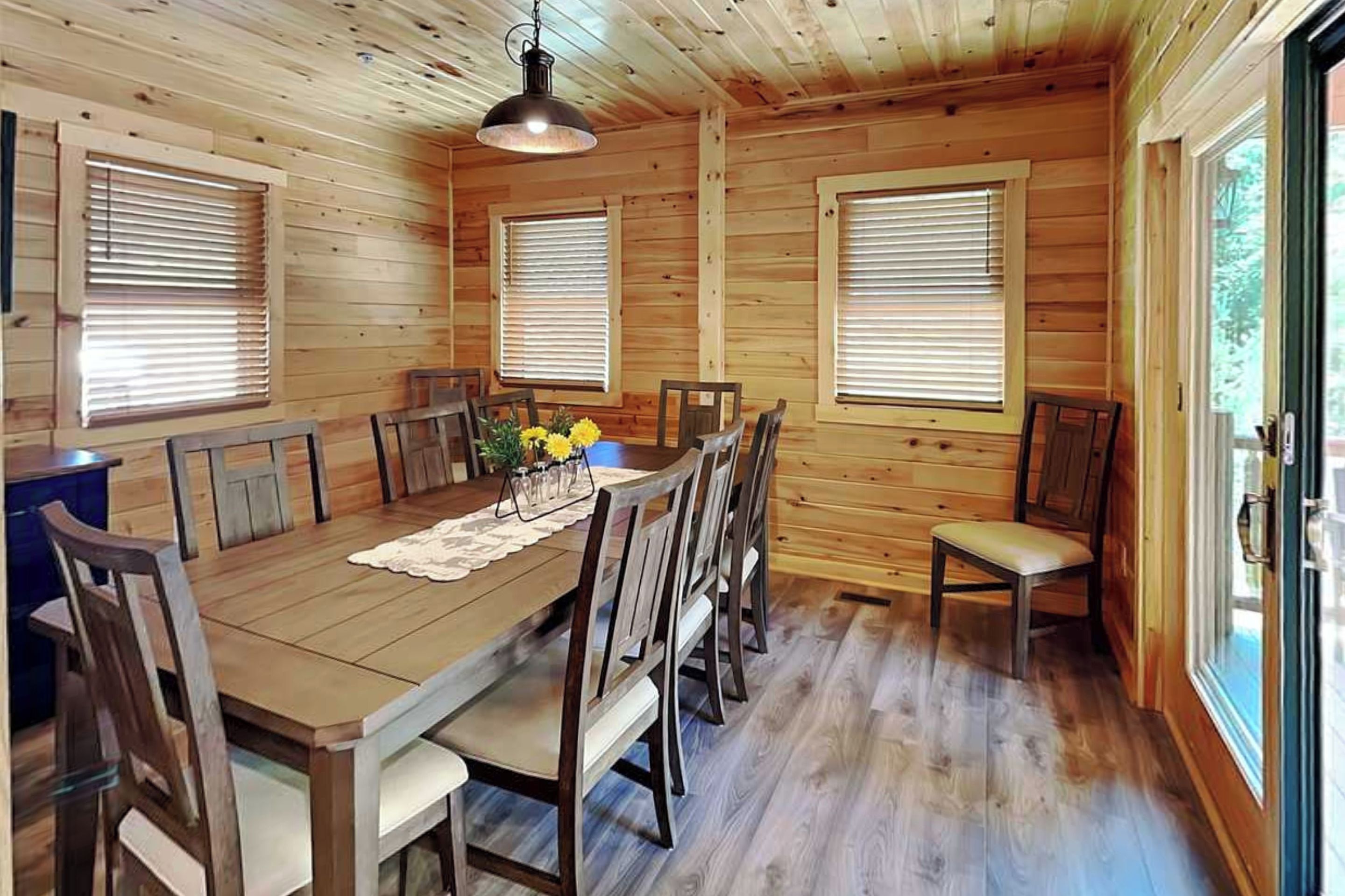 A wooden dining room with a table set for six, flanked by windows with blinds, and a hanging light fixture above.