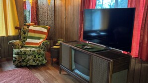 Tunnelmallinen olohuone, jossa on puupaneloitu seinä, televisio puujalustalla, kukkakuviollinen nojatuoli ja tähtimäinen riippuvalaisin.