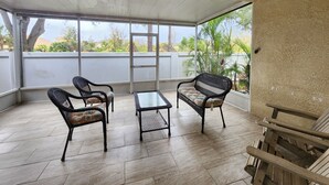 A covered patio area with two wicker chairs and a small table, large windows offering a view of greenery and buildings, and a ceiling fan.
