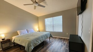 A bedroom with a bed, a ceiling fan, a nightstand with a lamp, a window with blinds, and a TV mounted on the wall.