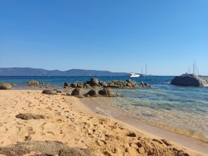 A sandy beach with clear water, large rocks, and boats in the distance.