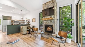 A modern kitchen with a stone fireplace, wooden furniture, and a large window overlooking greenery. - Sandhill - Luxury Cabin (Flathead County)