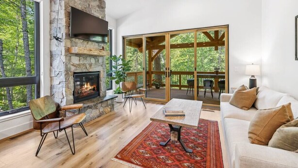 A spacious living room with a stone fireplace, a large window overlooking a deck, and a cozy seating area with a sofa and chairs. - Sandhill - Luxury Cabin (Flathead County)