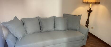 A light grey sofa with four cushions against a white wall, a wooden floor, and a lamp with a patterned lampshade.