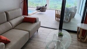 En moderne stue med sofa, sofabord i glas og udsigt til et bjergrigt landskab gennem store glasdøre.