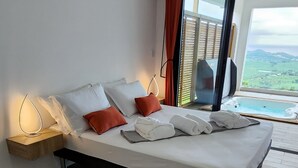 Et moderne soveværelse med trægulv, en seng med hvide og orange puder, et natbord med en lampe og et stort vindue med udsigt over landskabet.