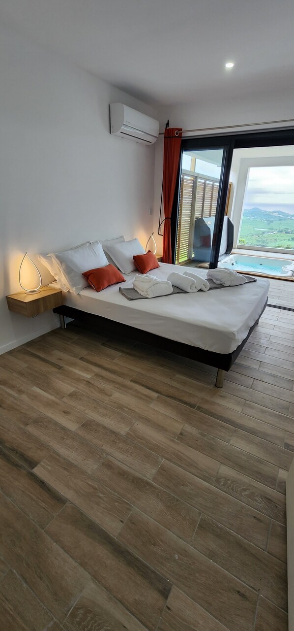 A modern bedroom with a wooden floor, a bed with white and orange pillows, a bedside table with a lamp, and a large window with a view of the landscape.