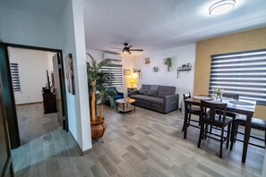 Ein modernes Wohnzimmer mit einem grauen Sofa, einem Esstisch mit Stühlen, einem Deckenventilator und einer großen Kühlpflanze.