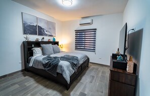 Ein Schlafzimmer mit einem Bett, einem Fernseher, einem Fenster mit Jalousien und einer Wand-Klimaanlage.