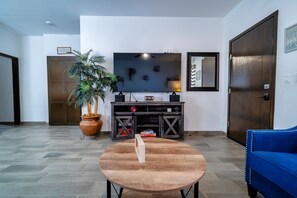 A living room with a round table, a blue sofa, a potted plant, a TV on a wooden stand, and a door with a sign.