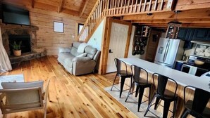 A cozy cabin interior with a wooden floor, a sofa, a dining area with chairs, a kitchen with a refrigerator, and a fireplace.