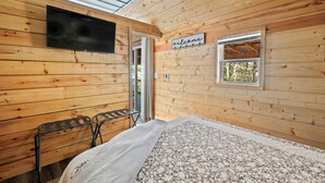 Ein Zimmer in einer Holzhütte mit einem Bett, einem an der Wand montierten Fernseher, einem Fenster mit einem 'welcome'-Schild und einem kleinen Tisch mit einem Stuhl.