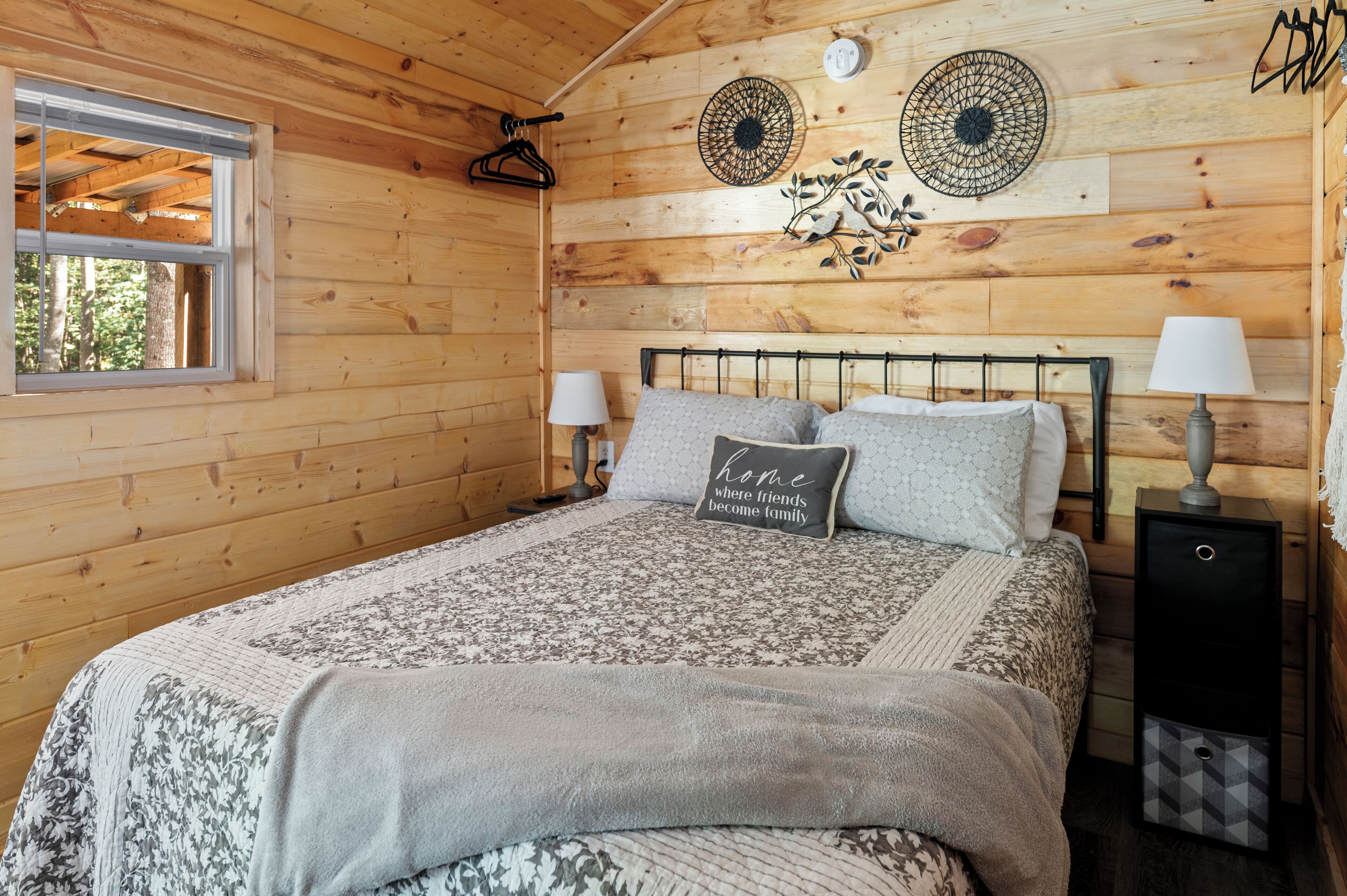 A wooden cabin bedroom with a bed, two wall-mounted lamps, a bedside table, and a window.