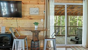 Ein Holzblockhaus mit Fernseher an der Wand, einem runden Holztisch mit zwei Metallstühlen, einem kleinen Topf mit Pflanze und einer Glastür zu einer Außenterrasse.