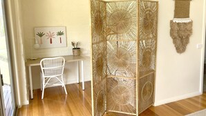 Quarto com piso de madeira, uma escrivaninha branca com cadeira, uma planta fixada na parede e um painel decorativo trançado.