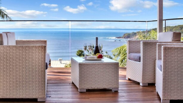 A balcony with wicker furniture, a small table with a bottle and glasses, and a scenic ocean view.
