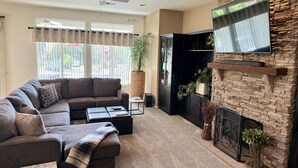 A living room with a sectional sofa, a flat-screen TV mounted on a stone wall, a fireplace, and a large window with curtains. - Casa Royal Troon- Active 55+ Golf Course Community, Private Home w Pool and Spa! (Indio)