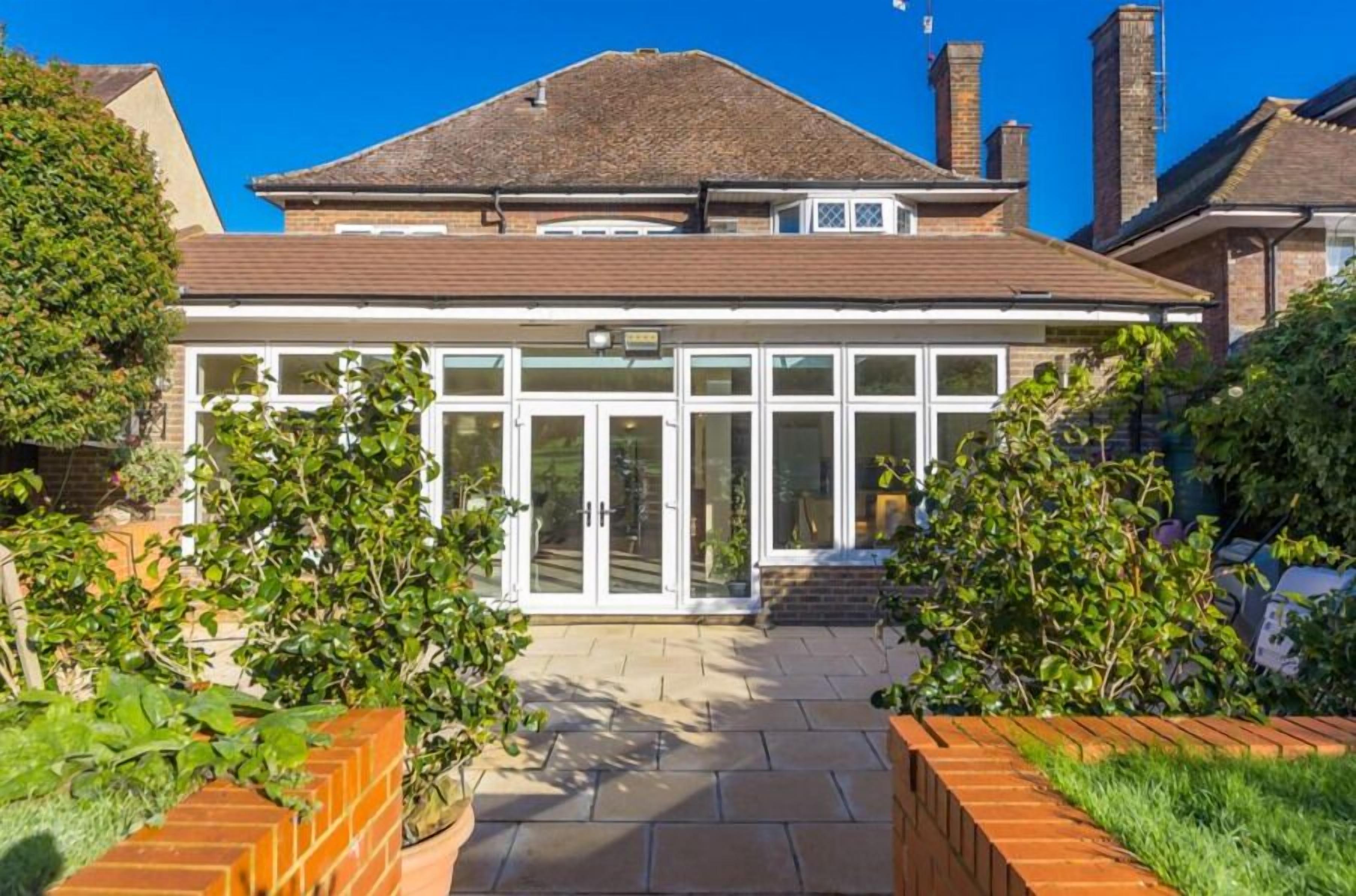 A house with a large glass door entrance, a brick pathway leading to the door, and a well-maintained garden with various plants.
