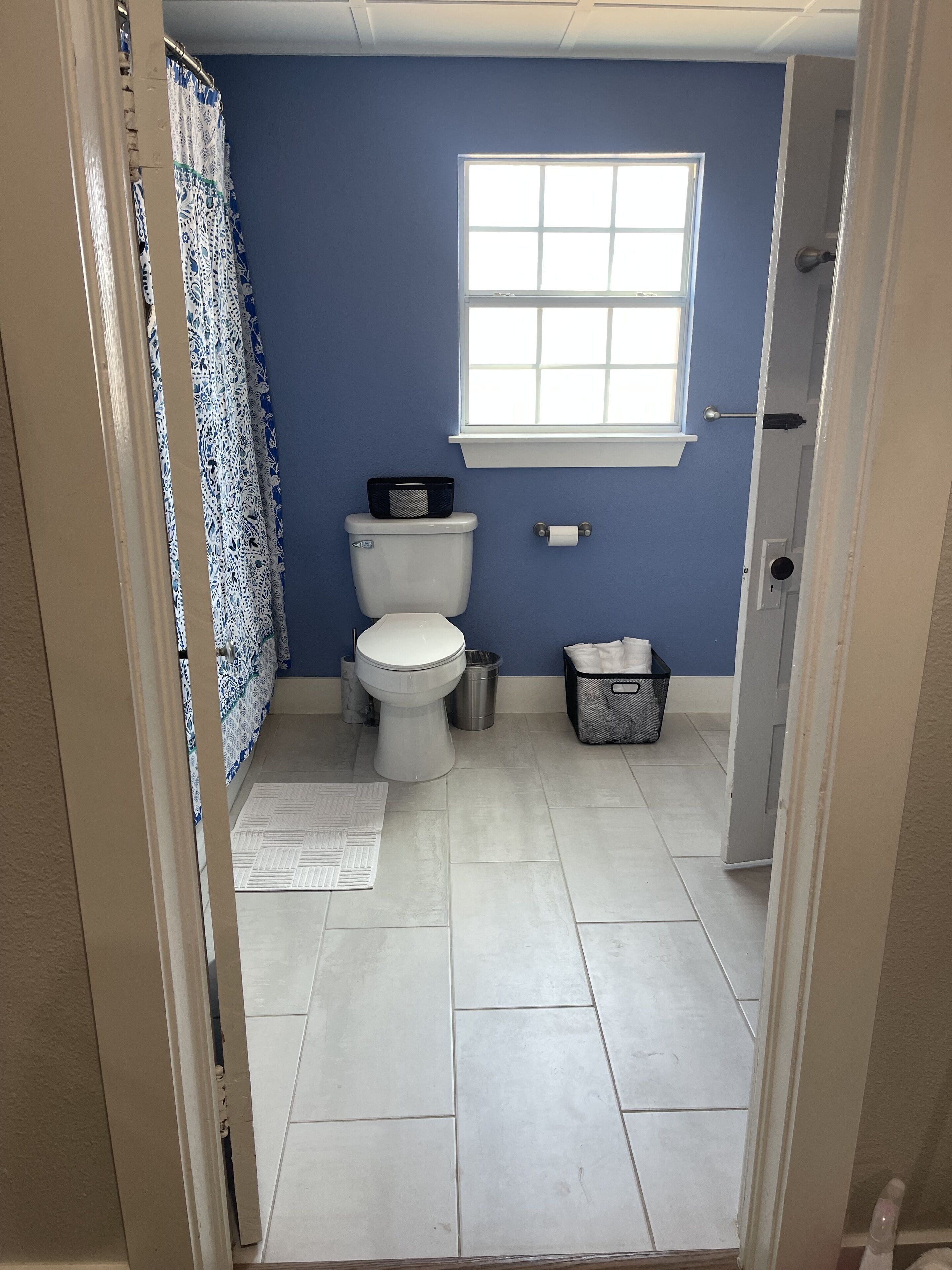 A bathroom with a toilet, a window, a shower curtain, a trash bin, and a storage basket.