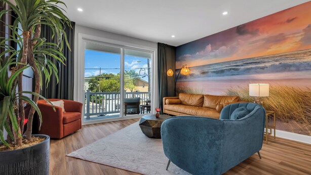 Ein Wohnzimmer mit großem Fenster, einer Couch, einem Sessel, einer Topfpflanze und einem Wandgemälde mit Strandmotiv.