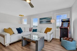 A modern living room with a sofa, a coffee table, and a foosball table. There is a window, a painting, and a small arcade machine.
