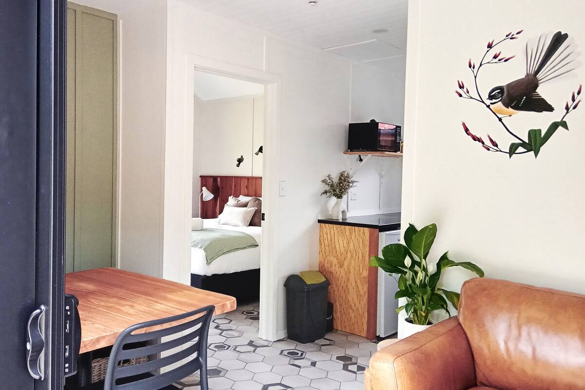 A modern apartment with a wooden dining table, a leather sofa, a kitchen area with a microwave, and a bed with a wooden headboard.