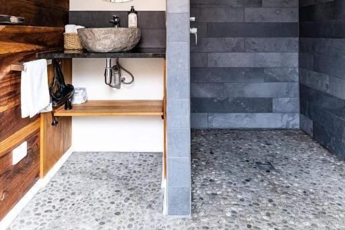 A modern bathroom with a wooden vanity, a round mirror, a stone sink, and a shower area with dark tiles.