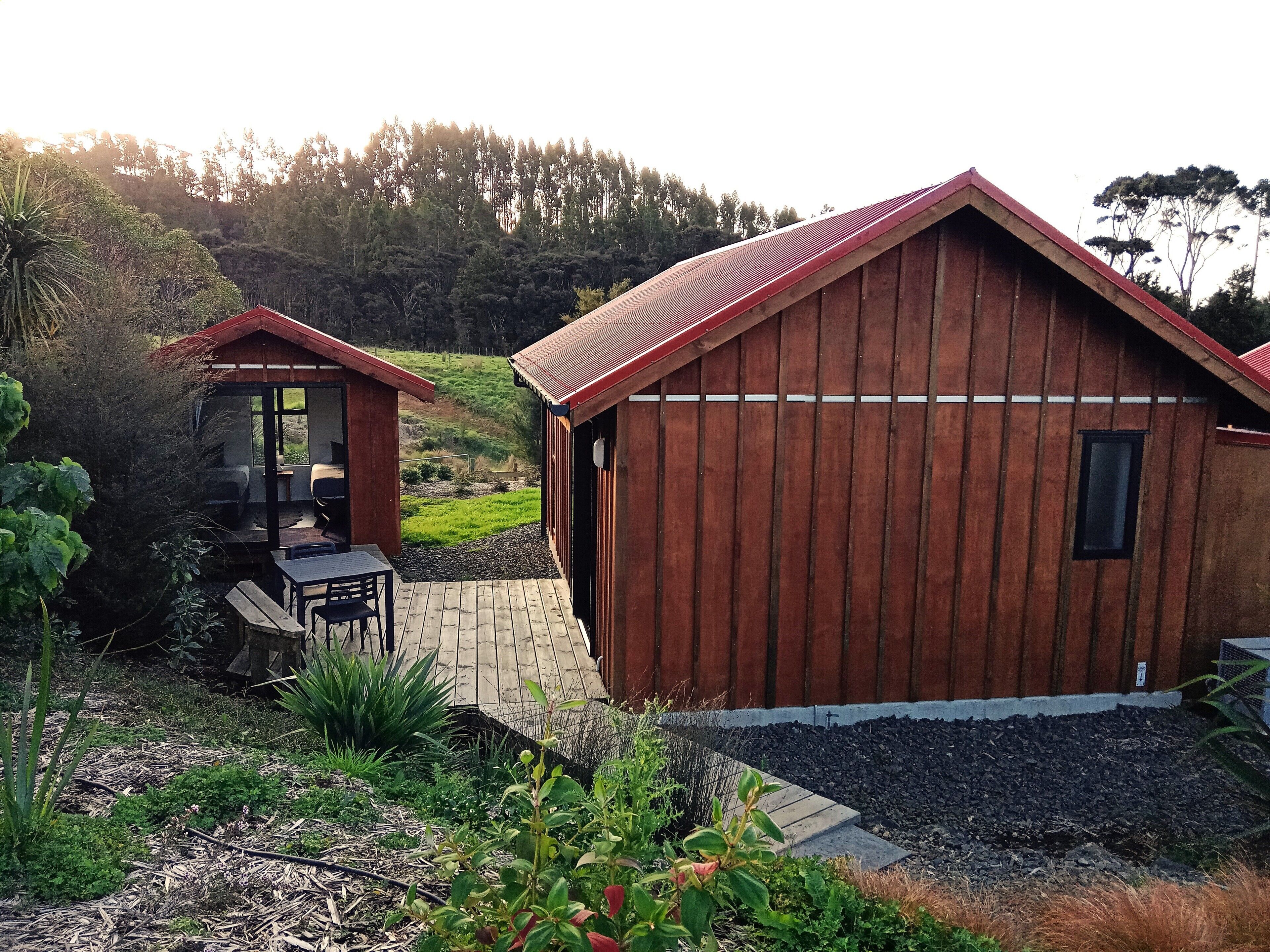 A wooden cabin with a sloped roof, a patio with a table and chairs, and a view of a green landscape.