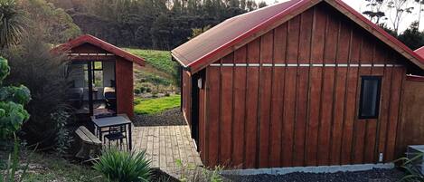A wooden cabin with a sloped roof, a patio with a table and chairs, and a view of a green landscape.