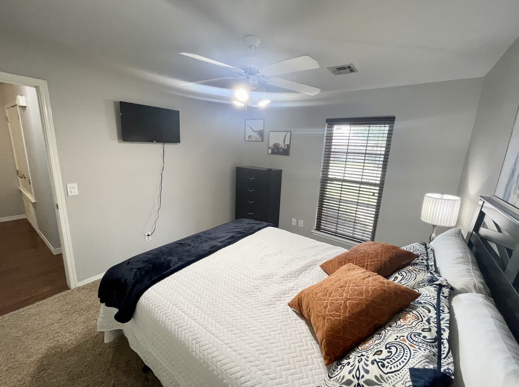 Une chambre à coucher comprenant un lit, un ventilateur de plafond, une télévision, une fenêtre avec des stores et une table de chevet avec une lampe.