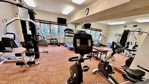 Un gimnasio con diverso equipo de ejercicio, incluyendo cintas de correr y máquinas de pesas, suelo alfombrado y ventanas amplias.