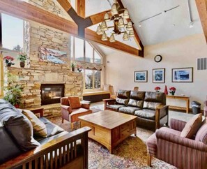 A spacious living room with a stone fireplace, leather sofas, wooden coffee table, and a large window providing natural light.