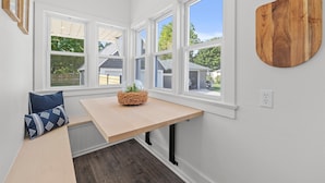 Et hjørneværelse med en træbænk, et lille bord og et vindue med udsigt til huse.