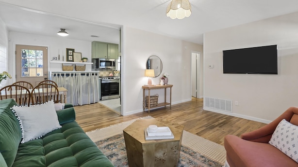 Een woonkamer met een groene bank, een houten salontafel en een roze fauteuil. Achtergrond is een keuken met een magnetron en een eettafel.