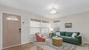 Een ruime woonkamer met houten vloeren, een groene bank, een roze fauteuil en een houten salontafel.