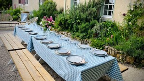 Mesa al aire libre con mantel a cuadros, platos y vasos, rodeada de follaje y un muro de piedra.