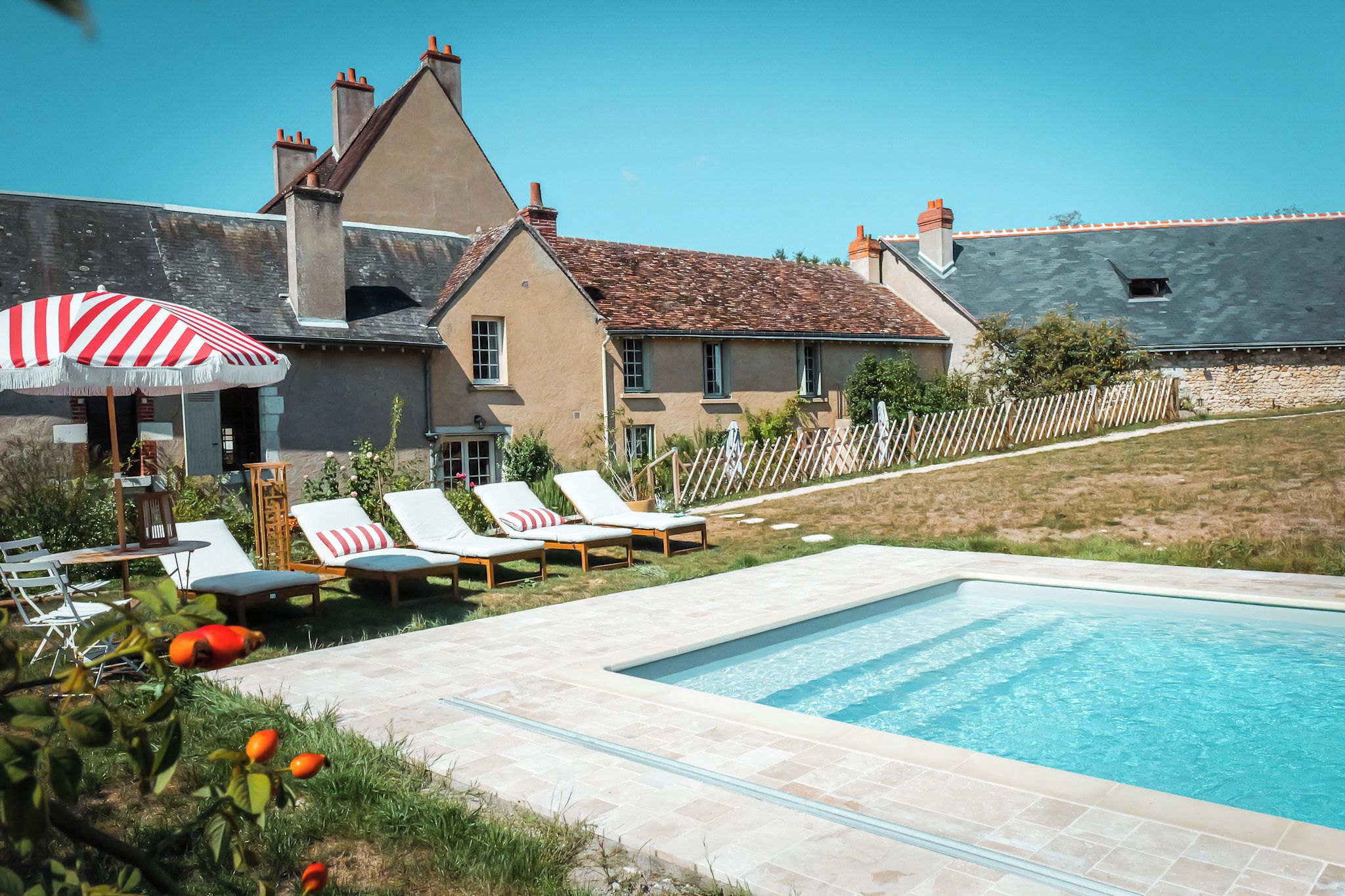 A swimming pool with lounge chairs and an umbrella, surrounded by a stone patio and a house with a tiled roof.